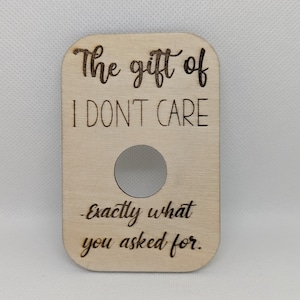 Op de afbeelding: Lichtgekleurde houten plaat met afgeronde hoeken en een cirkelvormige uitsparing. De tekst "The gift of I DON'T CARE" is boven de uitsparing geëtst, met "-Exactly what you asked for." eronder.