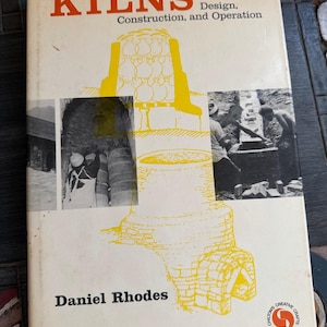 May include: A hardcover book titled KILNS Design, Construction, and Operation by Daniel Rhodes. The off-white cover features orange text, yellow line drawings of kilns, and two black and white photographs showing people working on kiln structures.