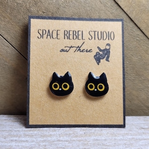 May include: A pair of black cat-shaped stud earrings with yellow eyes on a brown card. The card has the text "SPACE REBEL STUDIO out there" and a small illustration. The earrings are displayed on a wooden surface.