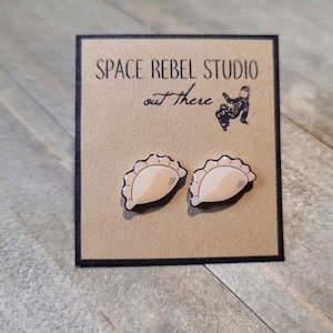May include: A pair of brown and white cartoon pierogi earrings on a brown card with the text "SPACE REBEL STUDIO out there".