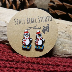 May include: A pair of penguin stud earrings with red and white striped scarves and blue feet. The earrings are on a brown card with the text "Space Rebel Studio out there".