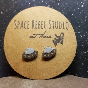 May include: A pair of silver and brown UFO stud earrings on a brown card with the text "SPACE REBEL! STUDIO out there".