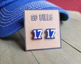 Puede incluir: Un par de pendientes esmaltados azules y blancos con el número 17, presentados en una tarjeta marrón con el texto "GO BILLS". Una gorra de béisbol azul está en el fondo. Un accesorio de temática deportiva.
