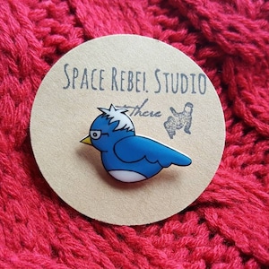 Può includere: Un uccello blu con accenti bianchi e un ciuffo di capelli bianchi sulla testa. L'uccello indossa gli occhiali e guarda a sinistra. L'uccello è su uno sfondo rotondo marrone con il testo "Space Rebel Studio" e "There" scritto su di esso.