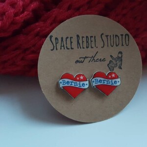 May include: A pair of red heart-shaped earrings with a white banner that says "Bernie" in blue lettering. The earrings are on a brown card with the text "Space Rebel Studio out there".