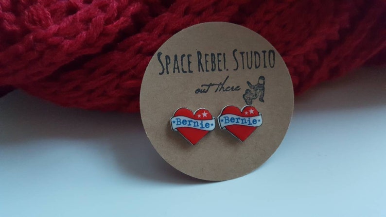 May include: A pair of red heart-shaped earrings with a white banner that says "Bernie" in blue lettering. The earrings are on a brown card with the text "Space Rebel Studio out there".