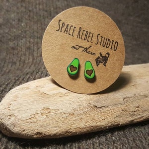 May include: A pair of avocado-shaped earrings with a brown heart in the center of each avocado. The earrings are on a small, brown card with the text "Space Rebel Studio out There" printed on it.