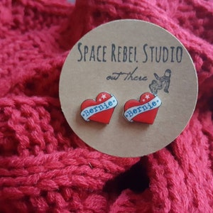 May include: A pair of heart-shaped earrings with the word "Bernie" printed on them. The earrings are red, white, and blue and are displayed on a brown card with the text "Space Rebel Studio out there".