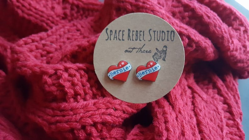 May include: A pair of heart-shaped earrings with the word "Bernie" printed on them. The earrings are red, white, and blue and are displayed on a brown card with the text "Space Rebel Studio out there".