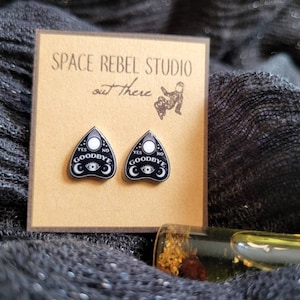 May include: A pair of black and white ouija board earrings with the words "YES NO GOODBYE" on them. The earrings are on a brown card with the text "SPACE REBEL STUDIO out there".
