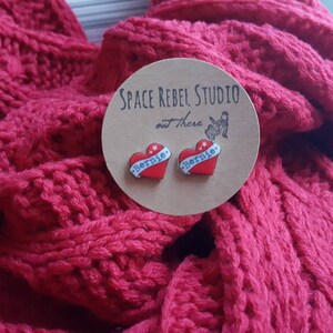May include: A pair of red heart-shaped earrings with the word "Bernie" written on them. The earrings are on a brown card with the text "Space Rebel Studio out there".
