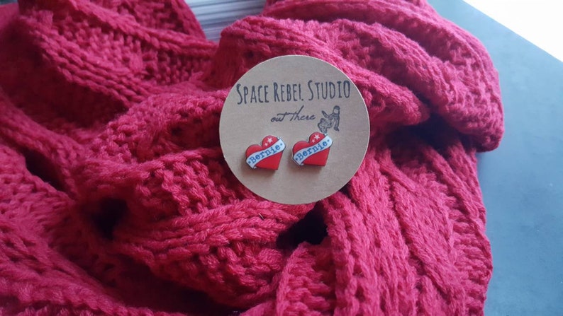 May include: A pair of red heart-shaped earrings with the word "Bernie" written on them. The earrings are on a brown card with the text "Space Rebel Studio out there".