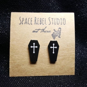 Könnte beinhalten: Ein Paar schwarze Sarg-Ohrringe mit weißen Kreuzen auf einer braunen Karte mit dem Text "Space Rebel Studio out There".