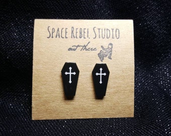 May include: A pair of black coffin-shaped earrings with white crosses on a brown card with the text "Space Rebel Studio out There".