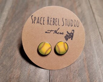 Peut inclure: Une paire de boucles d'oreilles clous avec un motif de balle de softball jaune et marron. Les boucles d'oreilles sont présentées sur une carte marron avec le texte "Space Rebel Studio" et un petit graphique d'astronaute.