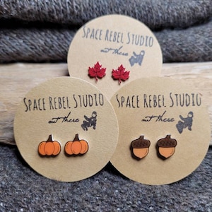 May include: Three sets of wooden earrings on brown cards. The cards have the text "Space Rebel Studio out there". The earrings are shaped like a maple leaf, a pumpkin, and an acorn.