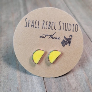 May include: A pair of yellow and green taco earrings on a brown card with the text "SPACE REBEL STUDIO out there".