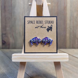 以下が含まれることがあります： 「Mafia Mafia」と書かれた紫と青のバイソン型のイヤリングのペア。イヤリングは、「Space Rebel Studio out there」と書かれた小さな木製イーゼルに飾られています。