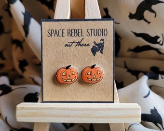 May include: A pair of orange pumpkin earrings with black eyes and a carved smile. The earrings are displayed on a small wooden easel with a brown card that reads "Space Rebel Studio out there".
