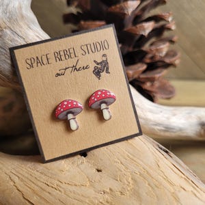 May include: A pair of red and white mushroom stud earrings. The earrings are on a brown card with the text "Space Rebel Studio out there".