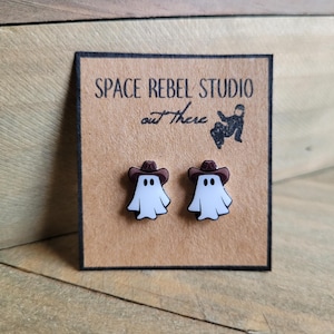 May include: A pair of white ghost earrings with brown cowboy hats. The earrings are displayed on a brown card with the text "SPACE REBEL STUDIO out There".
