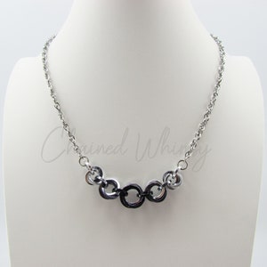May include: A silver-toned necklace with a chain and a series of connected rings. The central rings are black, while the others are silver. The necklace is displayed on a white mannequin bust.