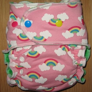 Può includere: Un pannolino di stoffa rosa con stampa arcobaleno e nuvole. Il pannolino ha un bordo verde e bottoni a pressione.