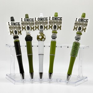 May include: Five decorative pens in a clear acrylic holder. Each pen has a unique design, including black glitter, olive green, white, and green with decorative accents. Each pen has a sign that reads "I ONCE PROTECTED HIM, PROUD ARMY, MOM."