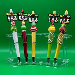 Puede incluir: Una colección de bolígrafos coloridos con el texto "Lab Tech" en la parte superior. Los bolígrafos tienen varios diseños, incluyendo barriles blancos, rojos, amarillos y verdes, y se exhiben en un soporte acrílico transparente sobre un fondo verde.