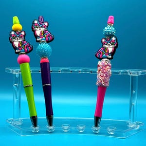May include: Three colorful pens with cat-themed toppers. The pens are green, purple, and pink. The cat toppers are pink, blue, and white with black stripes. The pens are displayed in a clear acrylic pen holder.