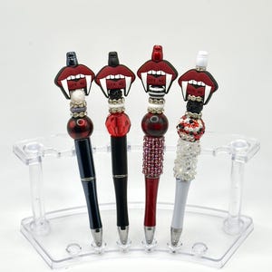 May include: Four decorative pens with red, black, and white accents. Each pen features a unique design, including a red mouth with the text "BITE ME". The pens are displayed in a clear plastic holder.