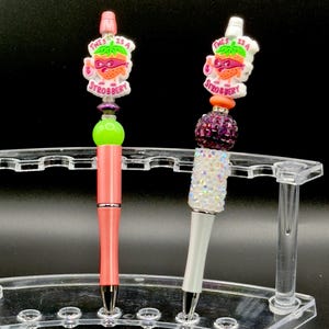 May include: Two pens with colorful strawberry-themed toppers. The toppers have the text "This is a Strawberry" on them. The pens are on a clear plastic stand.