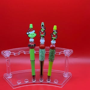 Op de afbeelding: Drie decoratieve pennen met Grinch-thema toppers, elk met een ander ontwerp. De pennen hebben groene en glitteraccenten en worden tentoongesteld in een doorzichtige acryl pennenhouder tegen een rode achtergrond. De Grinch-figuren dragen feestelijke kleding.