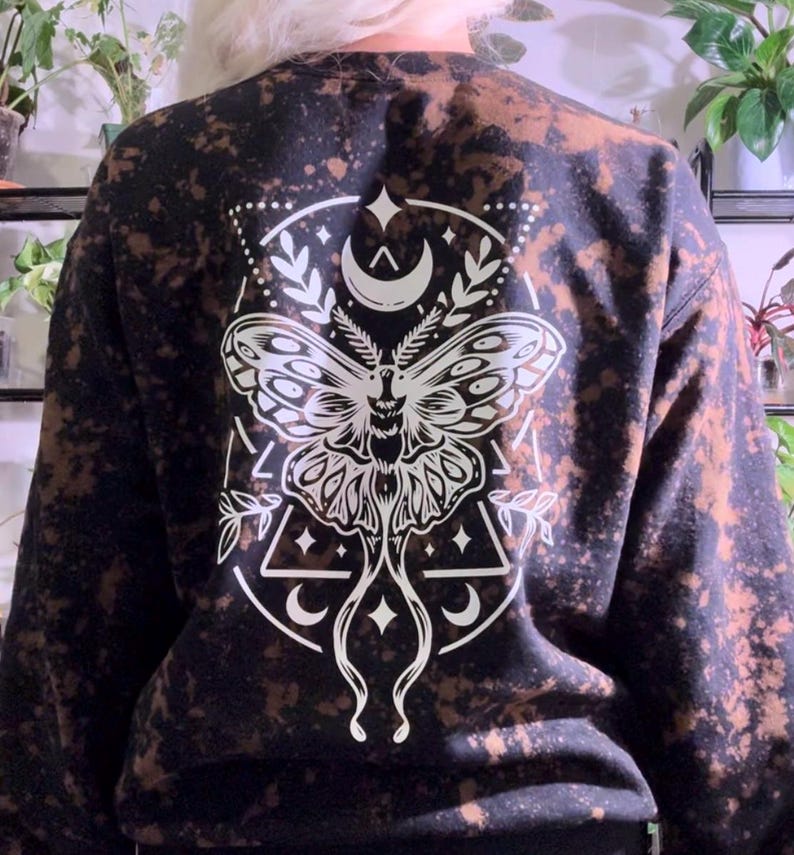 May include: Black sweatshirt with a bleached effect and a white graphic print of a moth with crescent moons and stars.