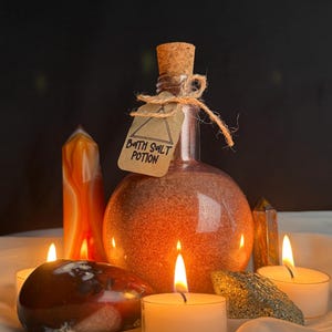 May include: A round glass bottle filled with orange bath salts, labeled "BATH SALT POTION", is sealed with a cork and twine. The bottle is surrounded by lit candles and crystals, creating a warm, inviting ambiance. The scene is set on a white cloth.