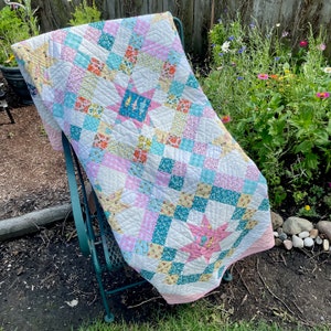 May include: A patchwork quilt with a star design in pink, blue, and yellow fabrics. The quilt is folded and draped over a green metal chair. The chair is sitting on a bed of brown mulch.