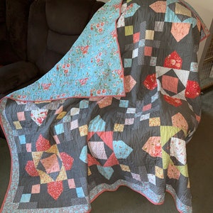 May include: A handmade quilt with a gray background and a patchwork design. The quilt features squares and diamond shapes in various colors, including pink, blue, and yellow. The reverse side is a light blue floral pattern. The quilt is trimmed with a red and white floral border.