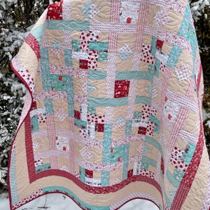 May include: A patchwork quilt in shades of beige, red, and teal. The quilt's design incorporates squares with strawberry, house, and other patterns. The border is a red and white floral pattern. The quilt is likely handmade.