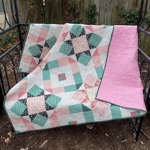 May include: A quilt with a patchwork design featuring pink, green, and black fabric. The quilt is folded and displayed on a metal bench.