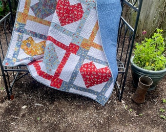 May include: A white quilt with a patchwork design featuring red, blue, and yellow hearts. The quilt is draped over a black metal garden bench.