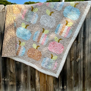 May include: A beige quilt with a patchwork design featuring various colored pumpkins. The pumpkins are in shades of blue, pink, brown, and white. The quilt is hanging on a wooden fence.