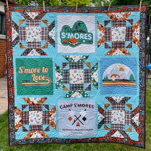 May include: A blue and brown patchwork quilt with a camp s'mores theme. The quilt features a variety of designs, including a campfire, a s'more, and the text "Camp S'mores".