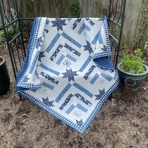 May include: A blue and white quilt with a star pattern. The quilt is folded and sitting on a wooden bench in a garden setting.