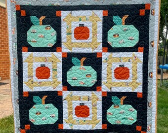 Puede incluir: Una colcha con fondo negro y un diseño de patchwork con manzanas verdes con autocaravanas blancas, calabazas naranjas y cuadrados blancos y amarillos.