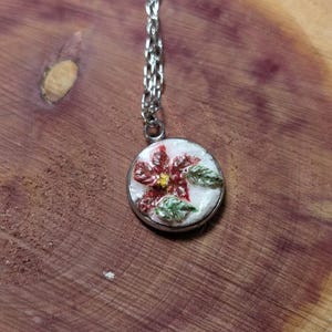 May include: A silver necklace with a round pendant featuring a red flower with green leaves on a white background. The pendant is set in a silver frame. The necklace is displayed on a wooden surface.