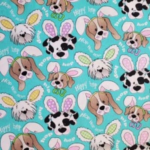 May include: A teal fabric featuring a repeating pattern of cartoon dogs wearing bunny ears. The dogs are brown, white, and black, with pink, green, and yellow accents. The words "Hop Hop" and "Hippy Hop" are scattered throughout the design.
