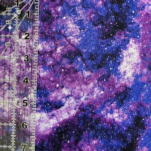 Puede incluir: Una tela con un vibrante dise&ntilde;o de galaxia. El patr&oacute;n presenta tonos arremolinados de morado, azul y blanco, salpicados de diminutos puntos blancos que parecen estrellas. Una regla est&aacute; colocada en el lado izquierdo de la tela.