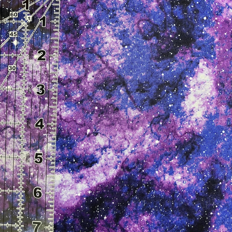 Puede incluir: Una tela con un vibrante dise&ntilde;o de galaxia. El patr&oacute;n presenta tonos arremolinados de morado, azul y blanco, salpicados de diminutos puntos blancos que parecen estrellas. Una regla est&aacute; colocada en el lado izquierdo de la tela.