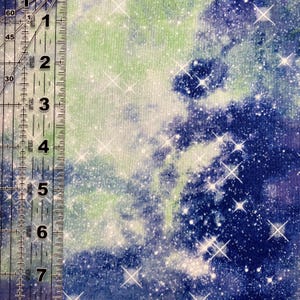 Puede incluir: Tela con un dise&ntilde;o celestial que mezcla tonos azules, verdes y blancos. La tela est&aacute; adornada con brillantes acentos en forma de estrella, creando un efecto c&oacute;smico. Una regla se coloca verticalmente a la izquierda, con medidas en cent&iacute;metros.