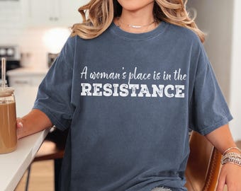 May include: A woman wearing a dark blue oversized t-shirt with the text "A woman's place is in the RESISTANCE" printed in white.
